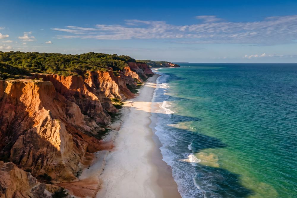 Fotografia aérea cinematográfica das falésias, da areia branca e do mar turquesa da Praia de Taípe, em Trancoso, Bahia, vista emoldurada por lotes no alto do relevo