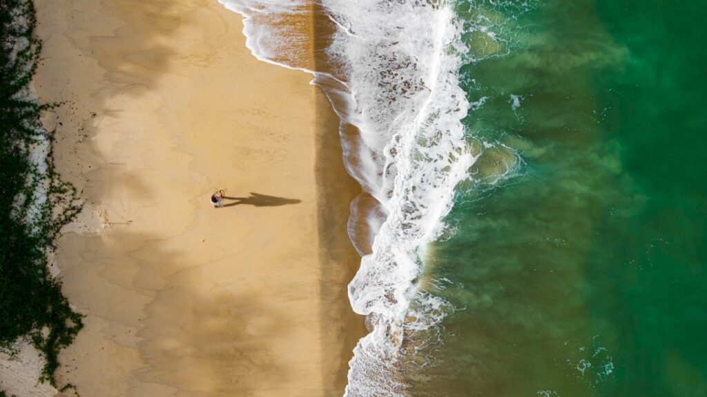 Faixa de areia de uma praia de Trancoso vista de cima, com uma pessoa caminhando à beira-mar, litoral da Bahia